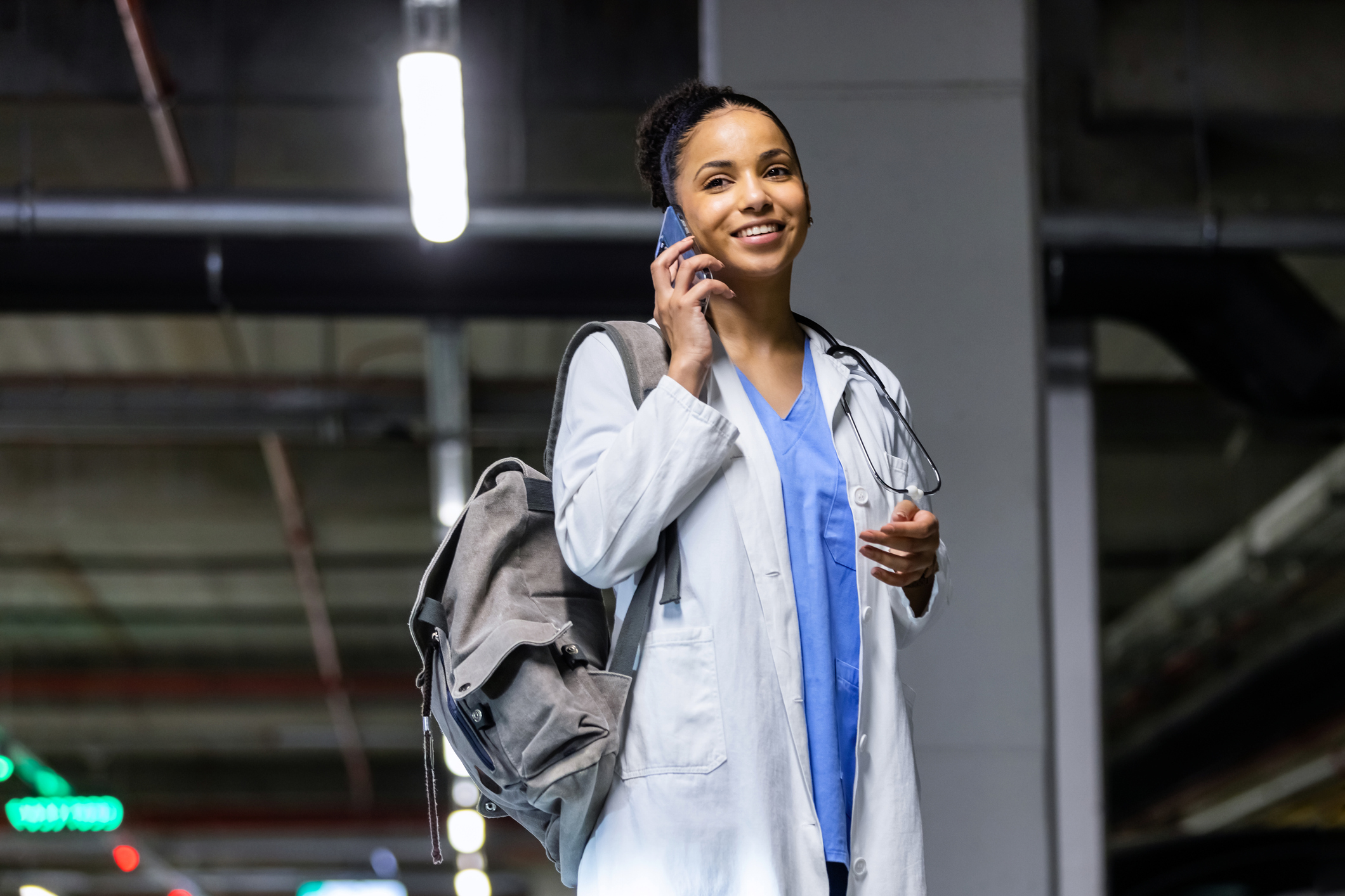 Confident, young female doctor walking along the parking lot while talking on the phone after having parked her car.