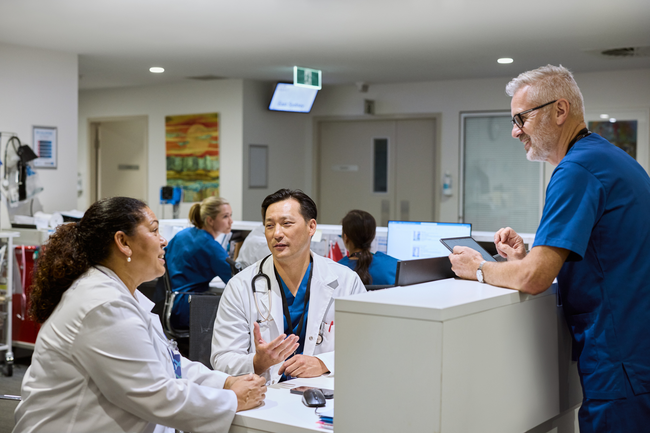Male doctor in white coat talking to colleagues at hospital workstation. Surgeon in blue scrubs holding digital tablet and listening. Healthcare professionals discussing patient care in medical office.