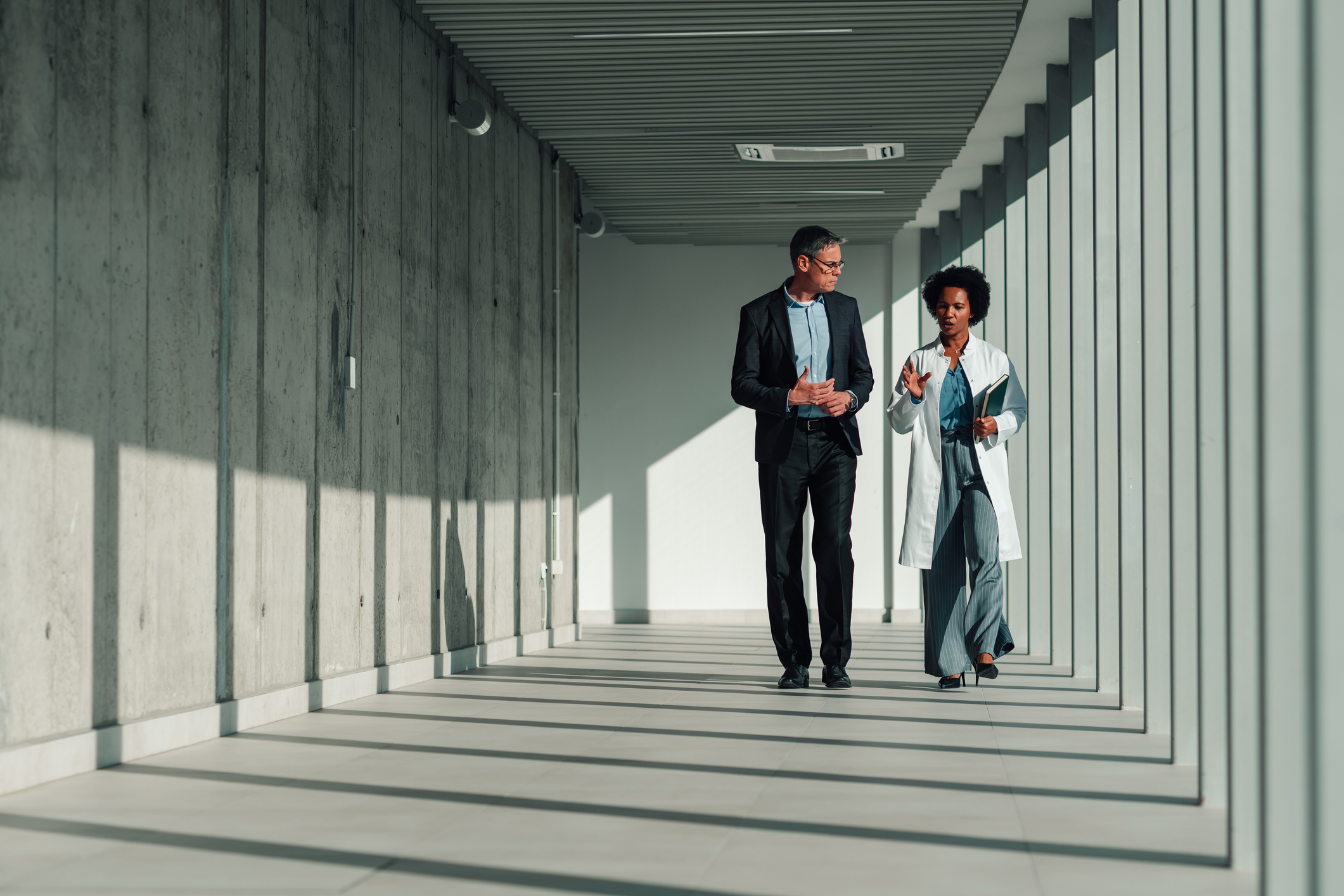 Diverse doctor and businessman walking through a bright, minimalist hospital corridor, having a serious professional discussion